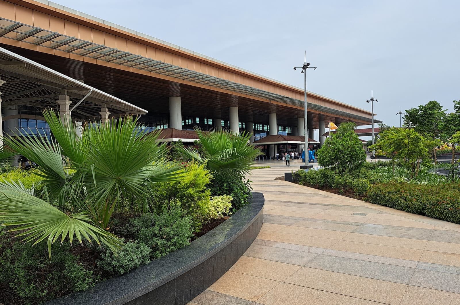 A paved walkway beside planted gardens leading to the columned terminal building at Mopa Airport