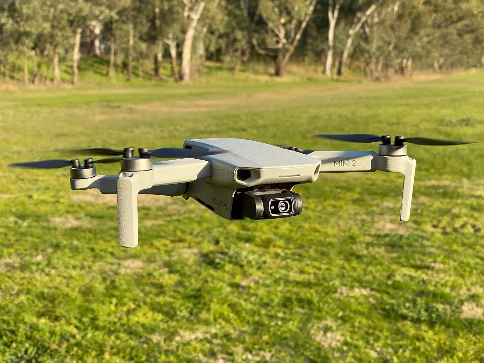 A small grey DJI drone flying low over a grassy field, with trees in the background