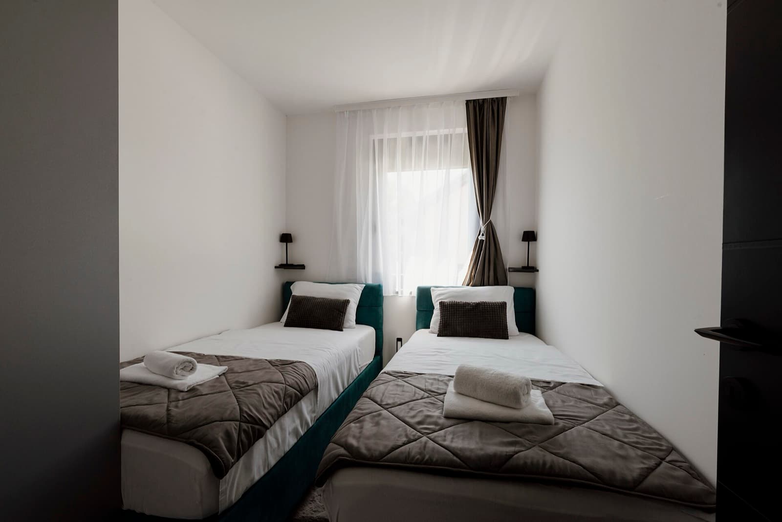 Two made single beds with grey quilts and rolled towels in a small white room with a curtained window