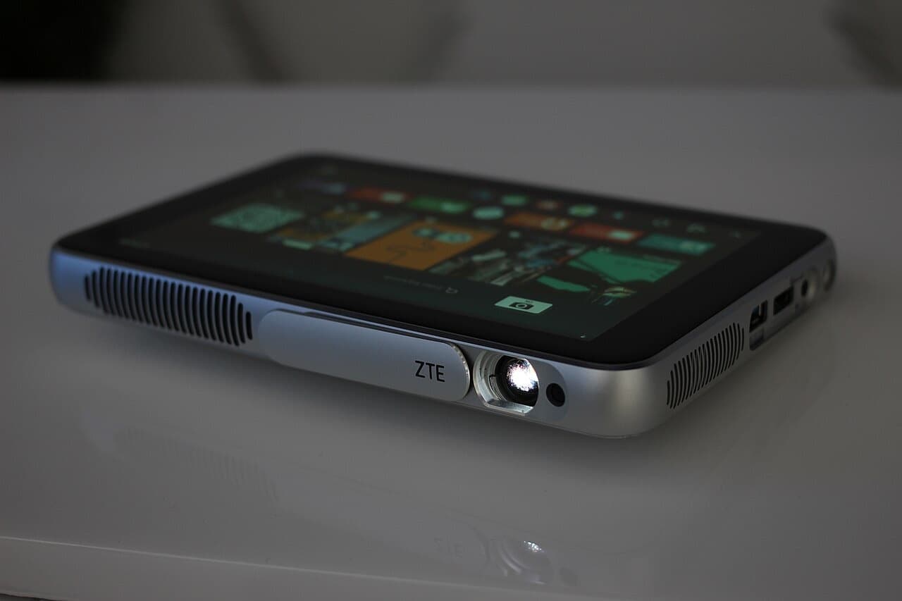 Compact smart projector on a table with its lens lit, showing side ports and speaker grilles