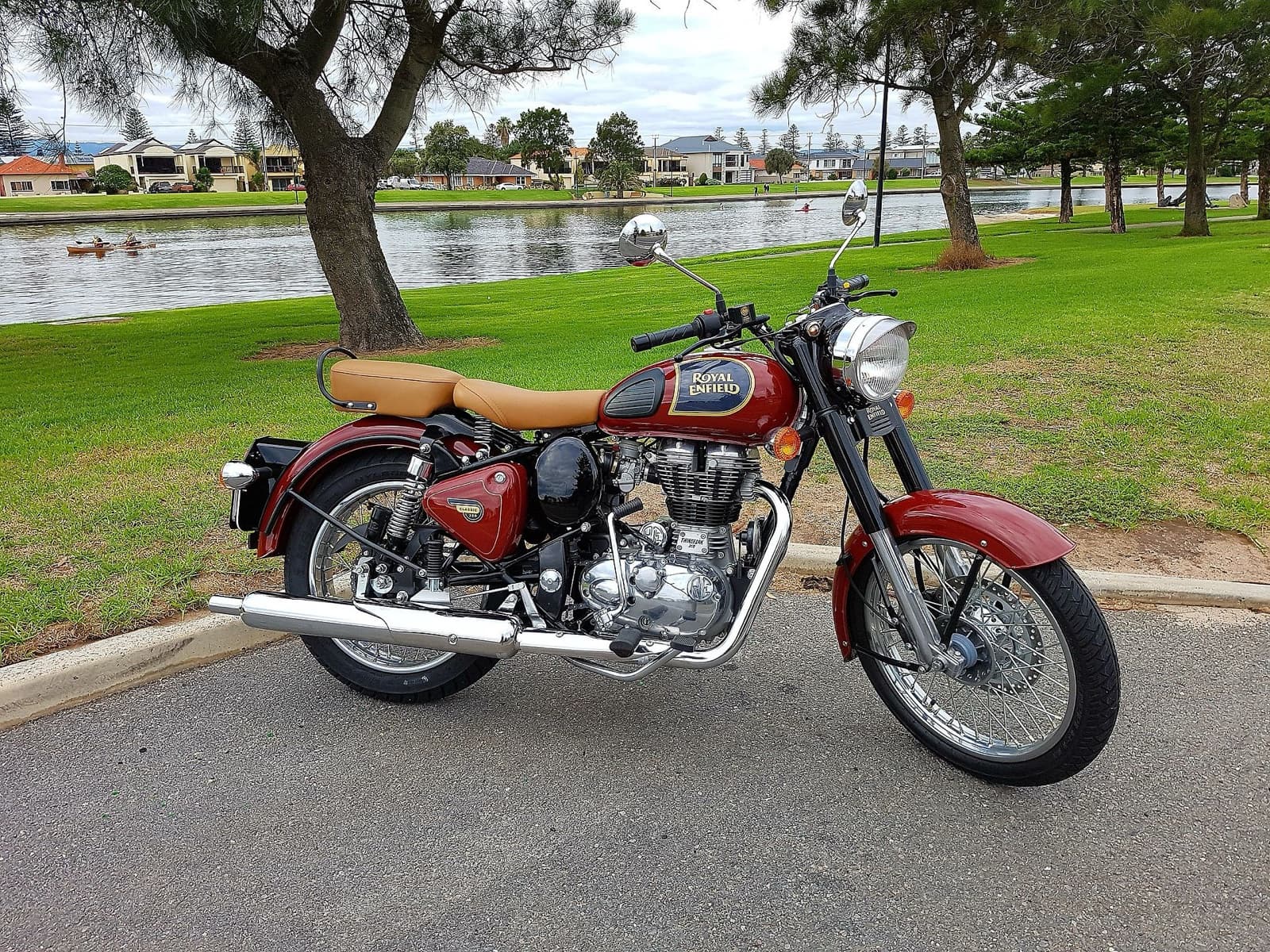 A maroon Royal Enfield motorcycle with a tan seat parked on a road beside a grassy lakeside park