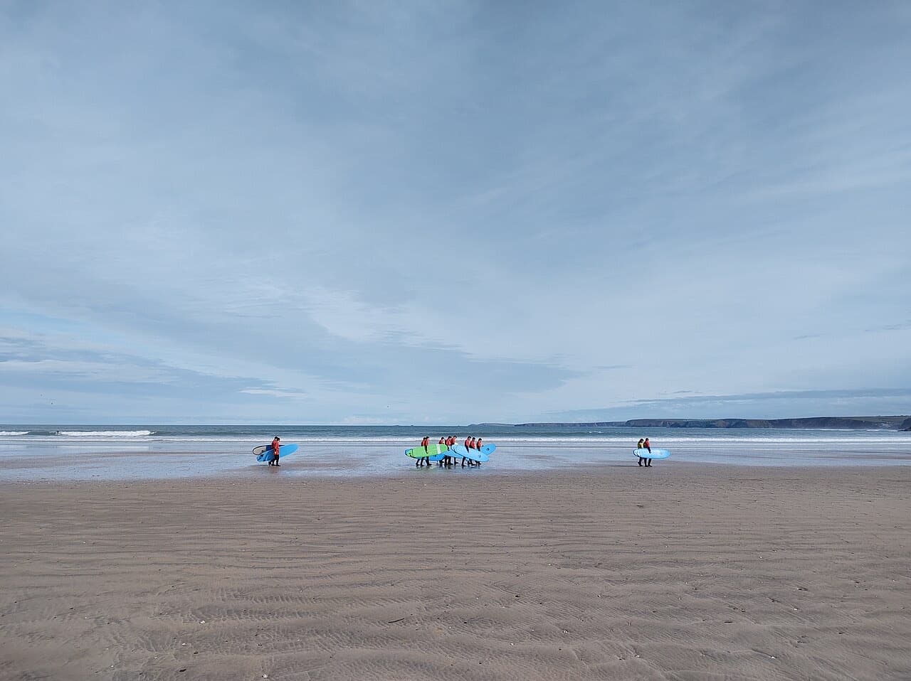 Beginner surfers carrying soft-top boards across a wide beach towards the waves