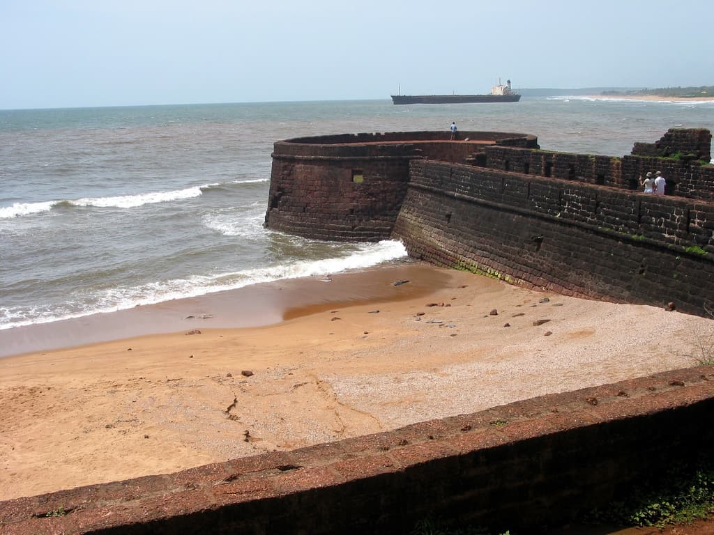 Laterite bastion of Fort Aguada curving into the sea beside a sandy cove, with a cargo ship on the horizon