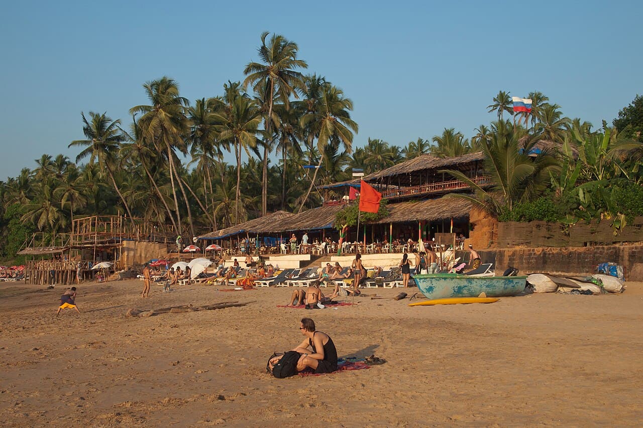 People relaxing on the sand at Anjuna Beach in front of Curlies beach shack, with palm trees and flags behind