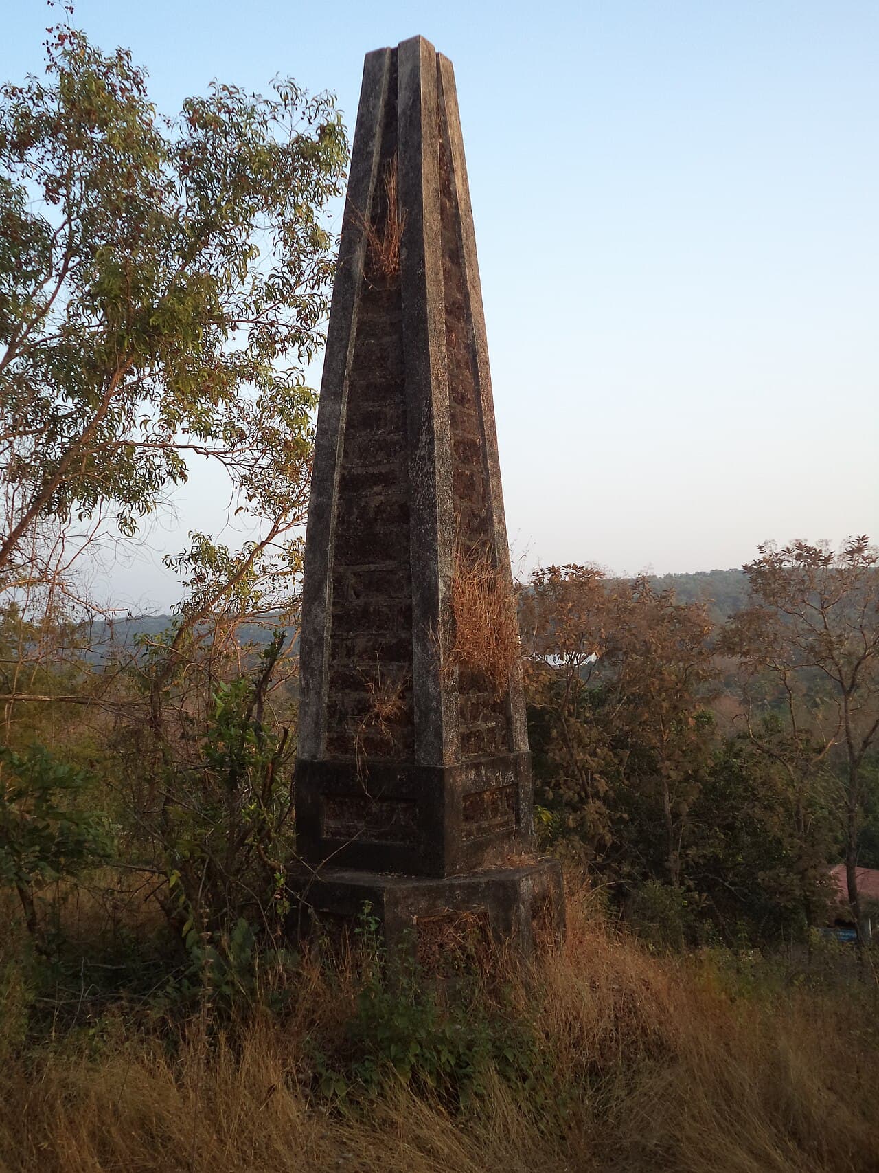 Weathered stone obelisk standing in dry grass on a hillside, with trees and hills behind it