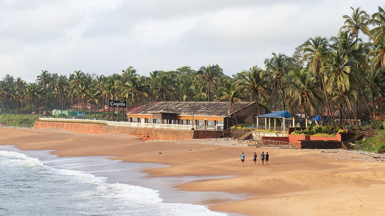 Waves reaching Sinquerim Beach as a few people walk on the sand past a low building and palm trees