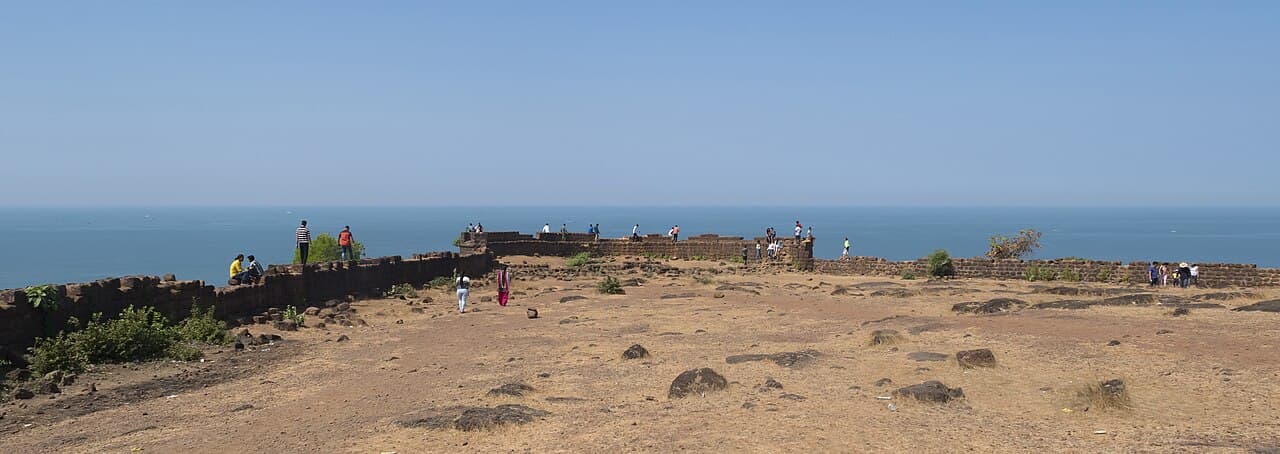 People walking along the laterite walls of Chapora Fort, with a dry rocky plateau and the open sea behind