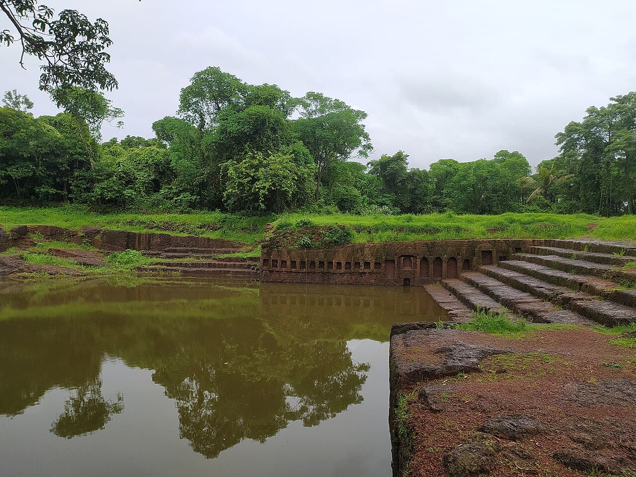 An old laterite temple tank on Divar Island with stone steps, still brown water and dense green trees