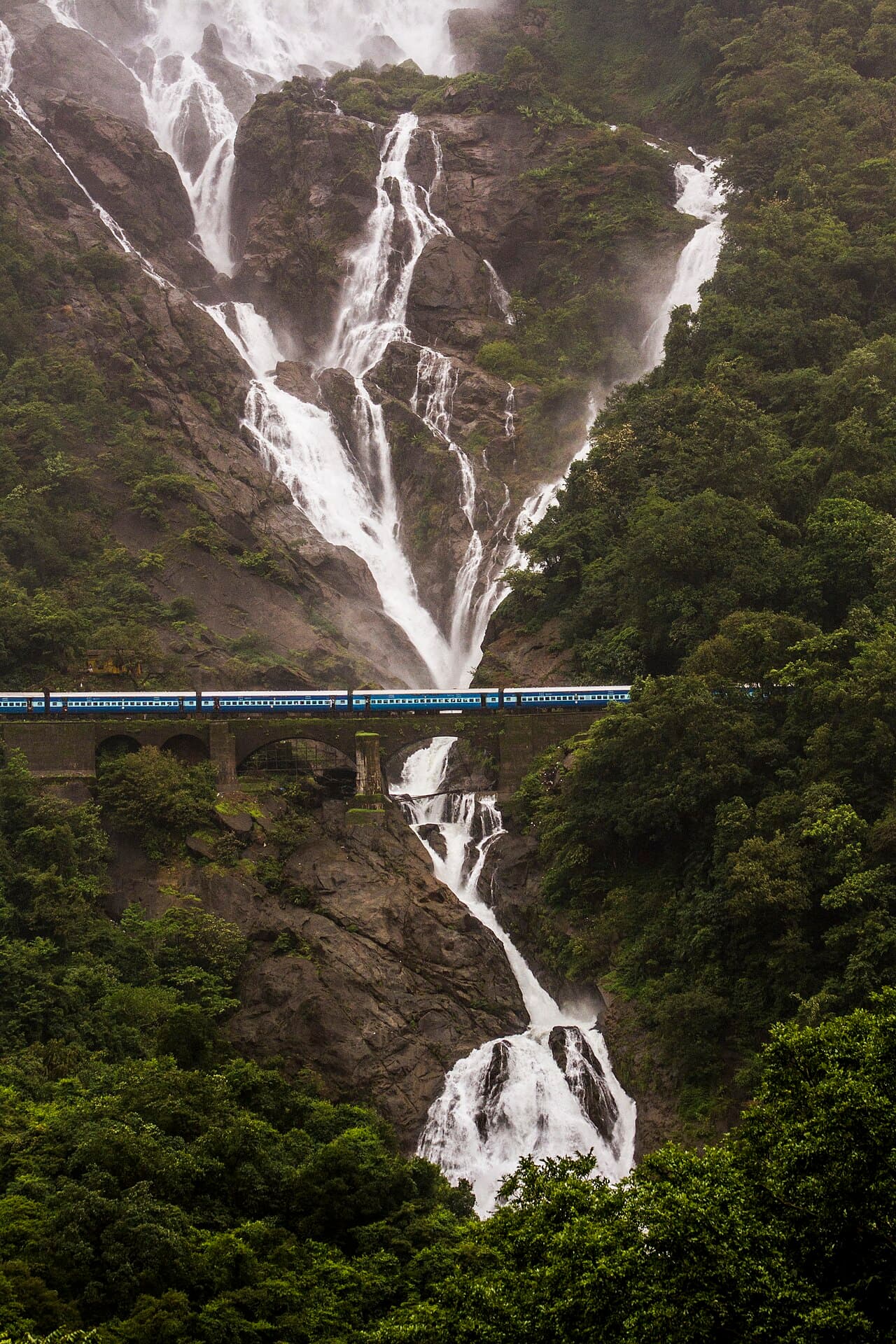 Dudhsagar waterfall pouring down a forested cliff, with a blue train crossing a stone bridge halfway down