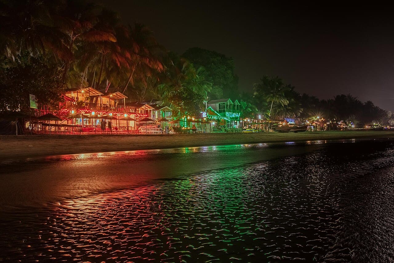 Beach huts glowing with red and green lights at Palolem Beach at night, reflected on the wet sand