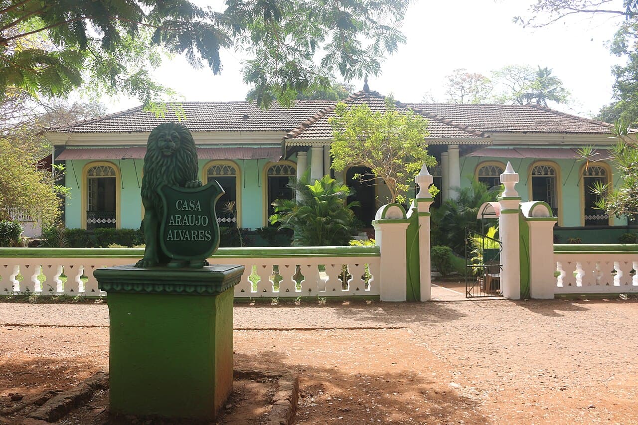 A mint green house with arched windows and a tiled roof behind a white wall, with a lion statue and nameplate