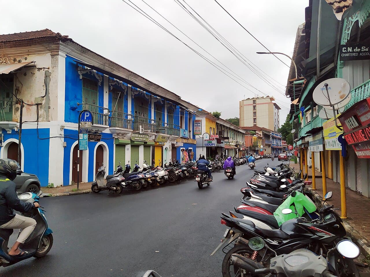 Street in Panaji's São Tomé quarter lined with parked scooters and blue colonial buildings, riders passing