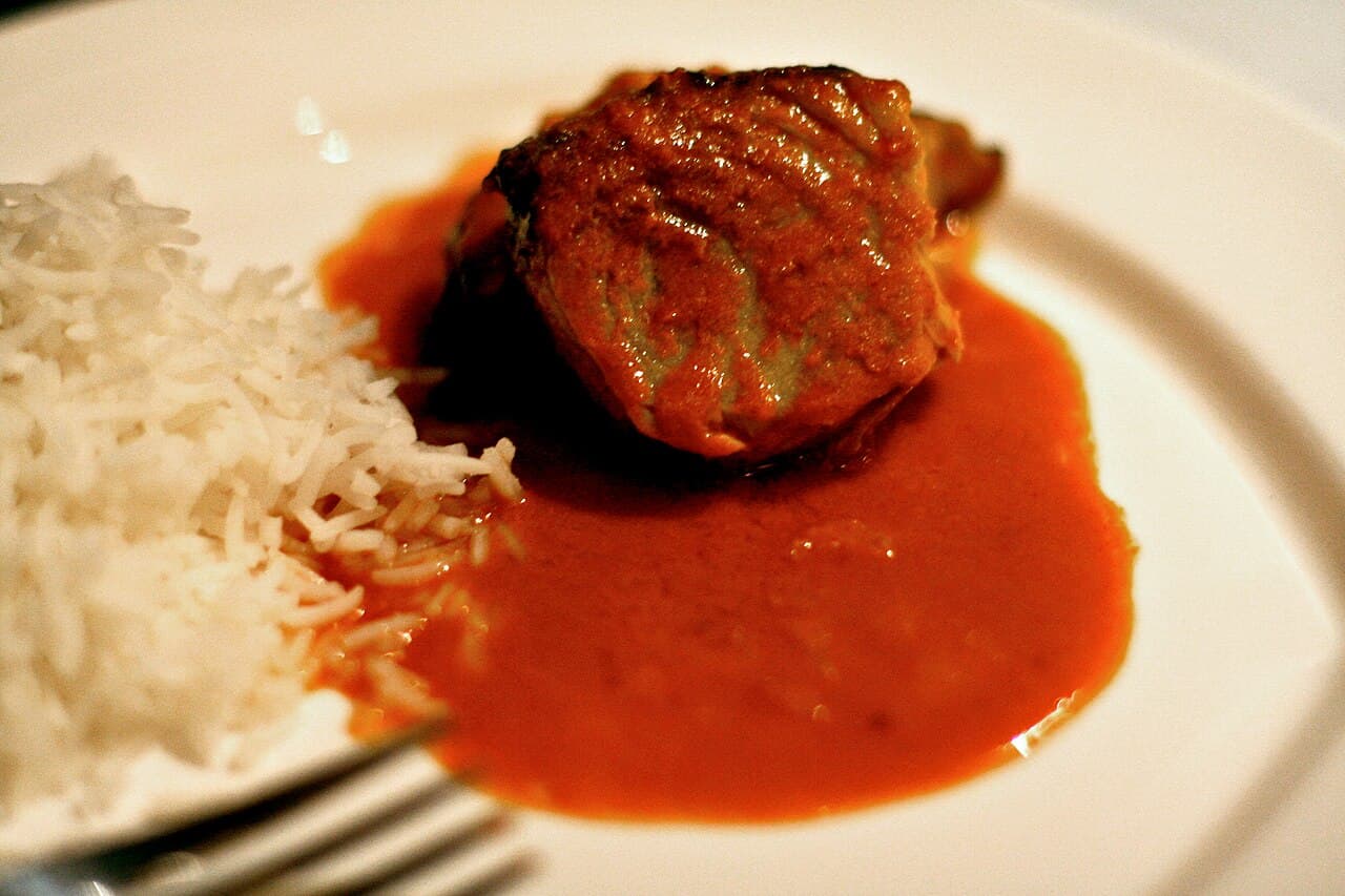 A piece of fish in red Goan curry beside a mound of white rice on a plate, with a fork in front