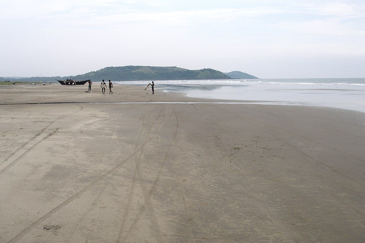 Wide, flat sand at Morjim Beach with tyre tracks, a few people walking and a green headland beyond